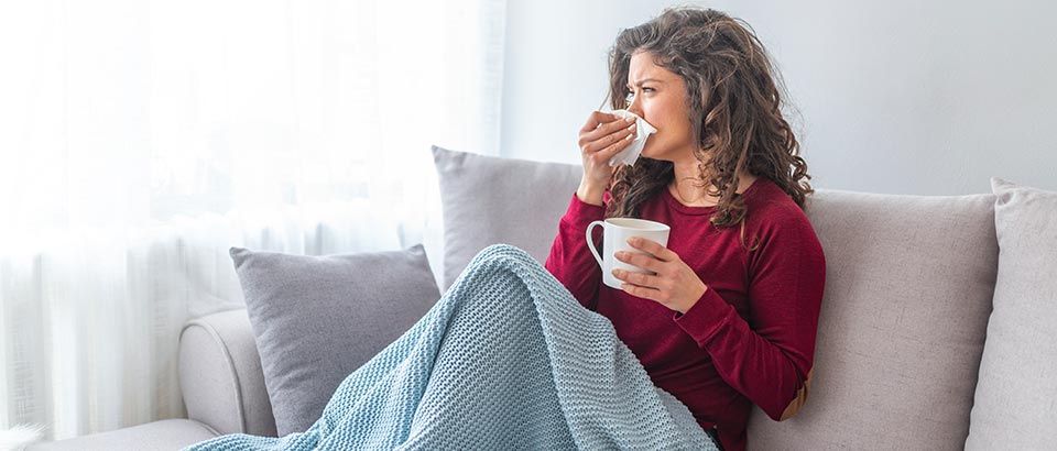 woman sitting on a couch holding a tissue to her nose and a cup of tea