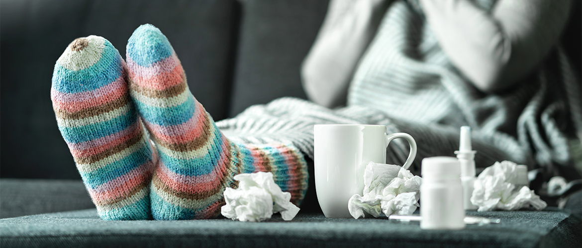 a sick person with their feet on a coffee table with used tissues and a tea cup sitting nearby