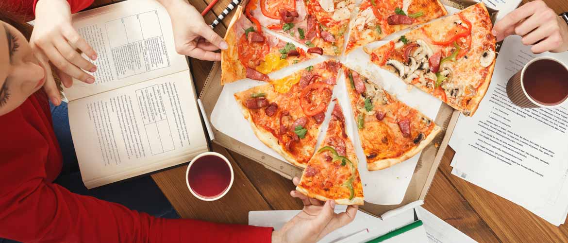 students studying and grabbing pieces of pizza from the center of a table that also contains notebooks and books