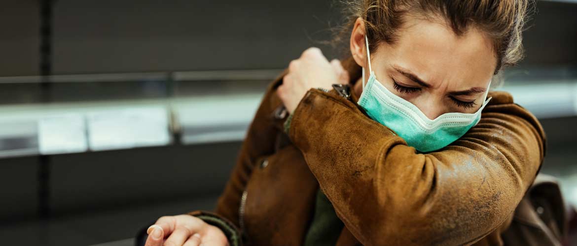 a woman wearing a mask and coughing into her arm while inside an office
