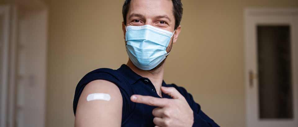 a man wearing a face mask and pointing to a bandage on his arm after receiving the COVID-19 vaccine