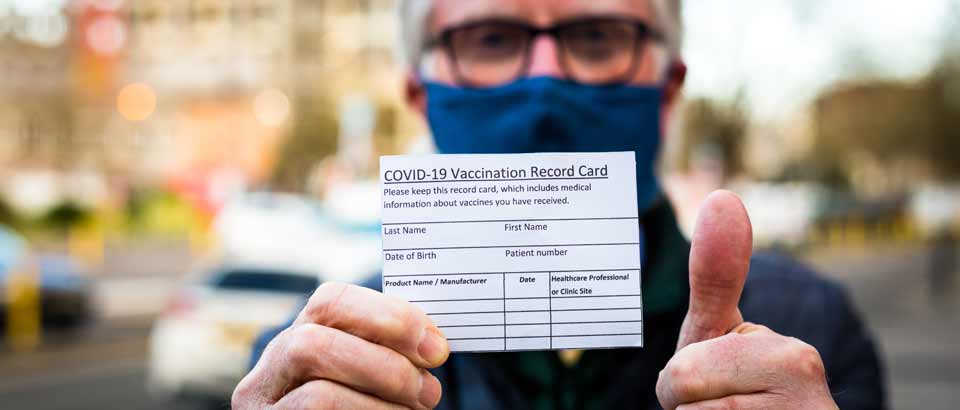 an older man wearing a face mask holding up his COVID-19 vaccination record card