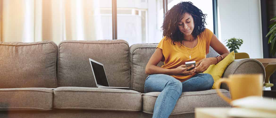 a woman sitting on a couch looking at her phone with a laptop sitting beside her