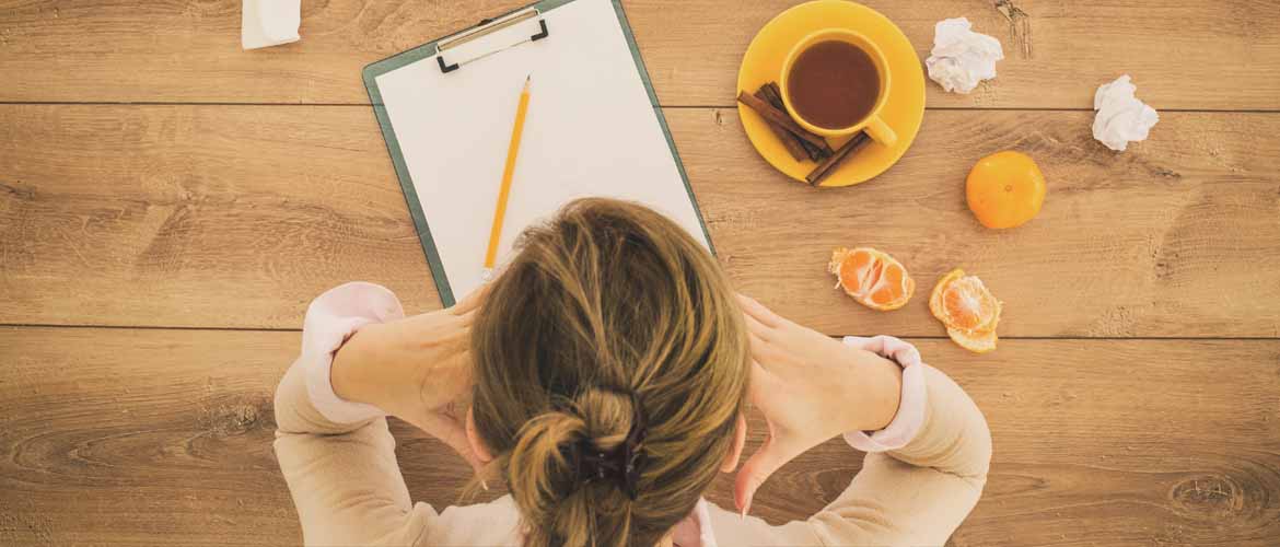 A woman not feeling well at a desk with a cup of tea, an orange and crumpled paper