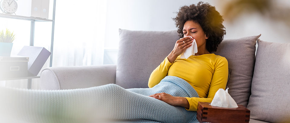 a woman sick with the flu sitting on a couch and holding a tissue against her nose