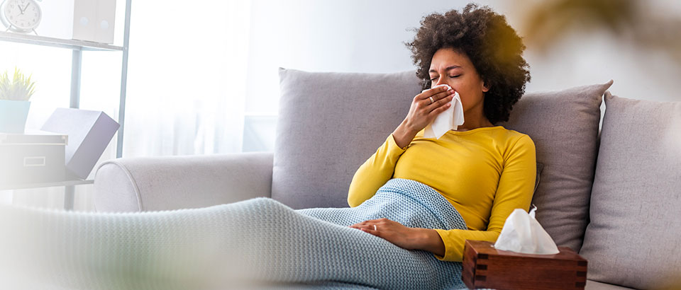 uma mulher doente com a gripe sentada num sofá e segurando um tecido contra o nariz