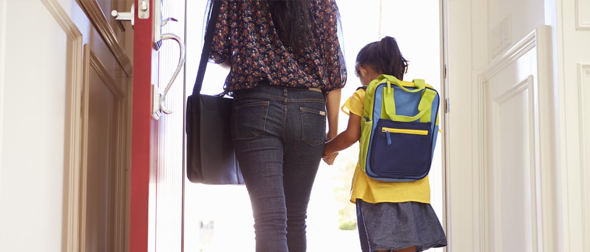 Rear view of a small school girl wearing a backpack holding her mom's hand and walking out the door