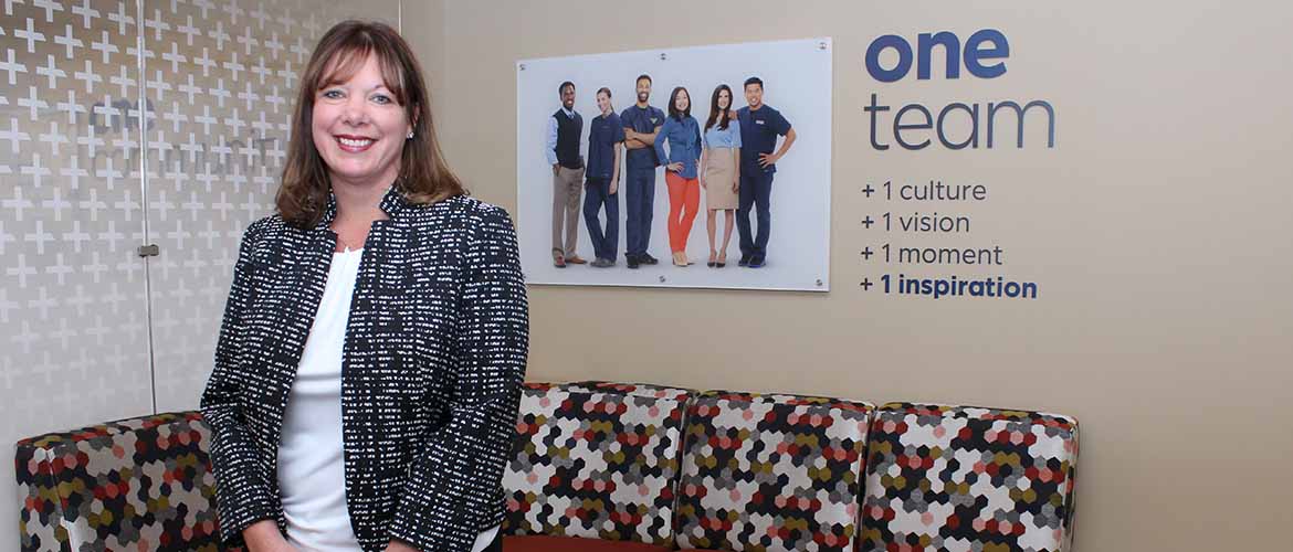 a photo of Amie Carter standing in front of a wall that reads, 'one team. one culture. one vision. one moment. one inspiration.