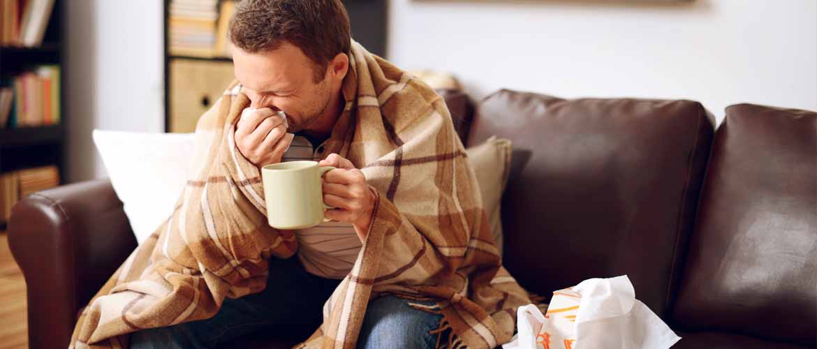 an ill man sitting on a couch and sneezing into a tissue