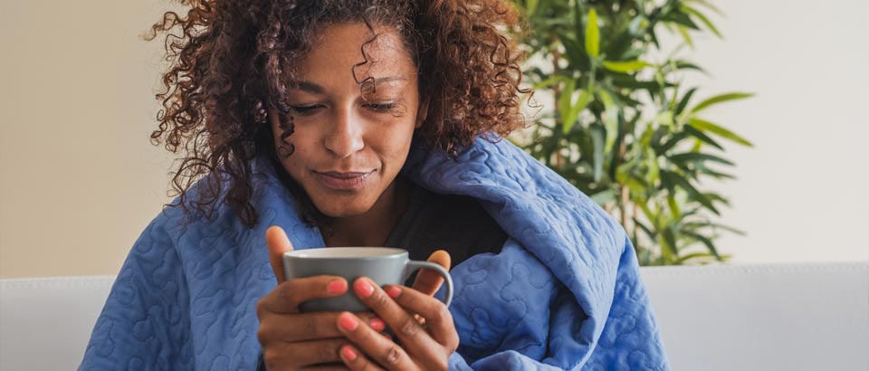 woman sick with blue blanket on shoulders holding a coffee mug